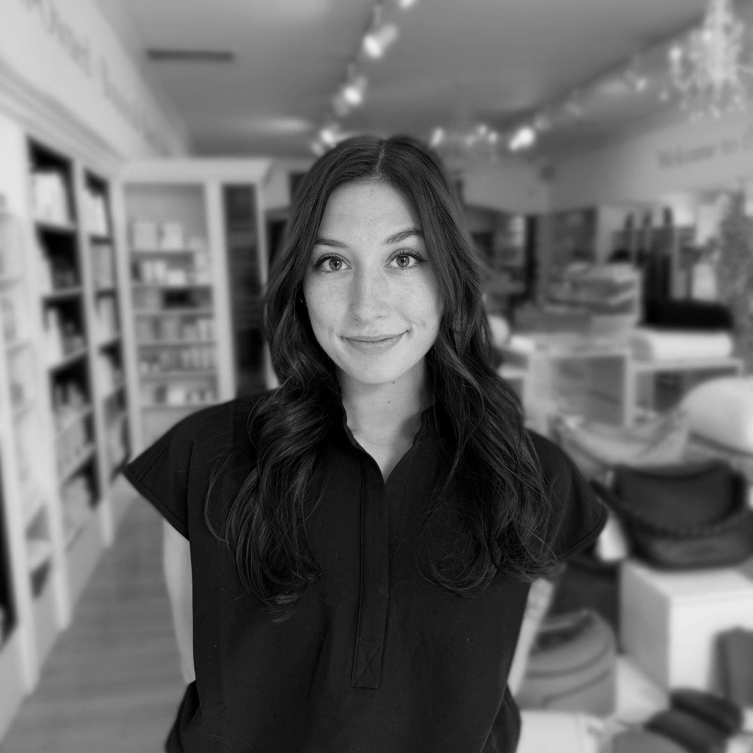 Black and white photo of a woman standing in a store with shelves and products in the background.