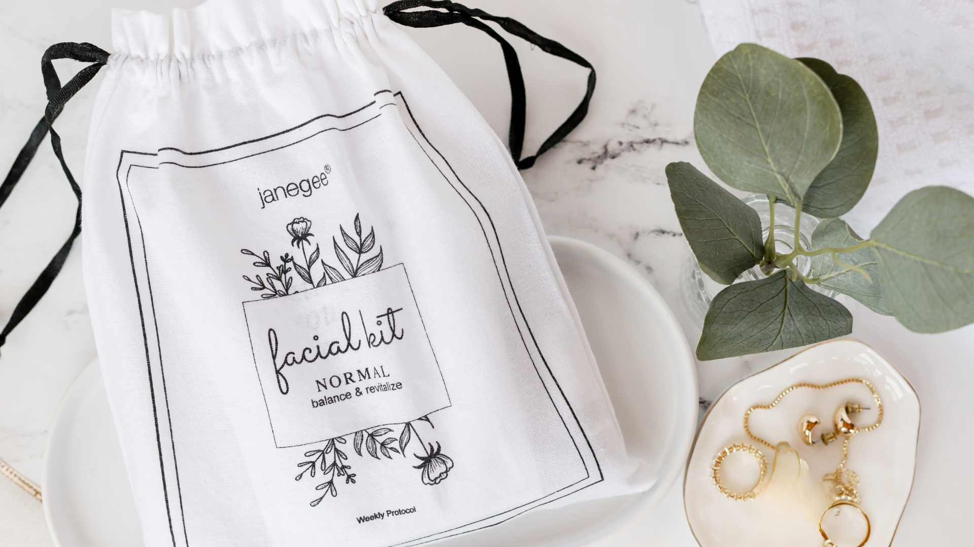 A white drawstring pouch with black ties and a printed label reading 'janeglow facial kit NORMAL balance & beauty' is styled on a white plate beside green eucalyptus leaves and gold jewelry on a light marble surface.