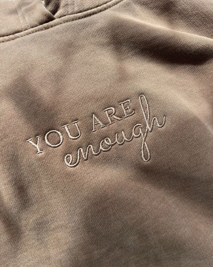 Close-up of the center chest embroidery on a brown janegee hoodie, showing the text "YOU ARE" in a block font with "enough" written in a cursive script underneath.