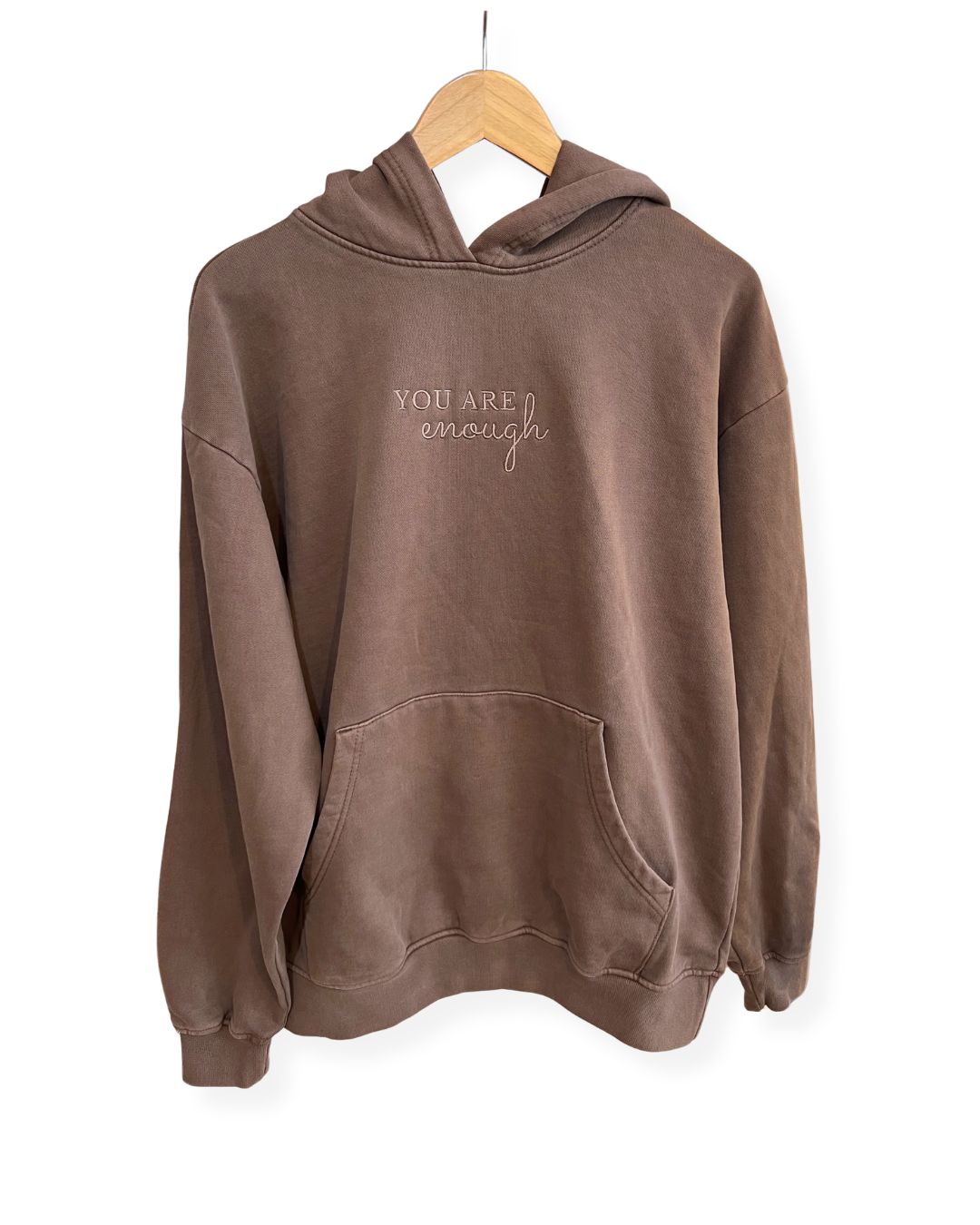 Front view of a brown, oversized fleece janegee hoodie hanging on a wooden hanger, featuring a kangaroo pocket and "YOU ARE enough" embroidered centered on the chest.