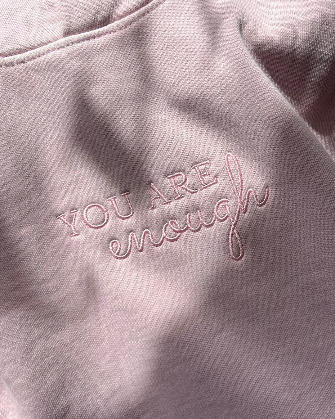 Close-up of the center chest embroidery on a light pink janegee hoodie, showing the text "YOU ARE" in a block font with "enough" written in a cursive script underneath.