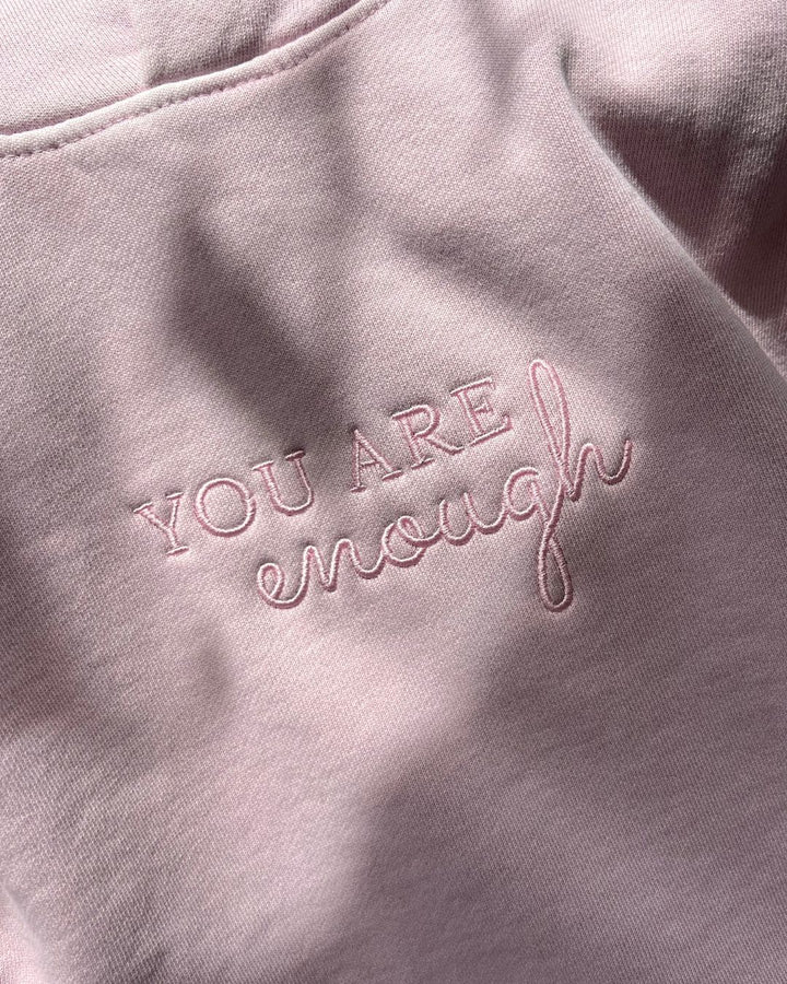 Close-up of the center chest embroidery on a light pink janegee hoodie, showing the text "YOU ARE" in a block font with "enough" written in a cursive script underneath.