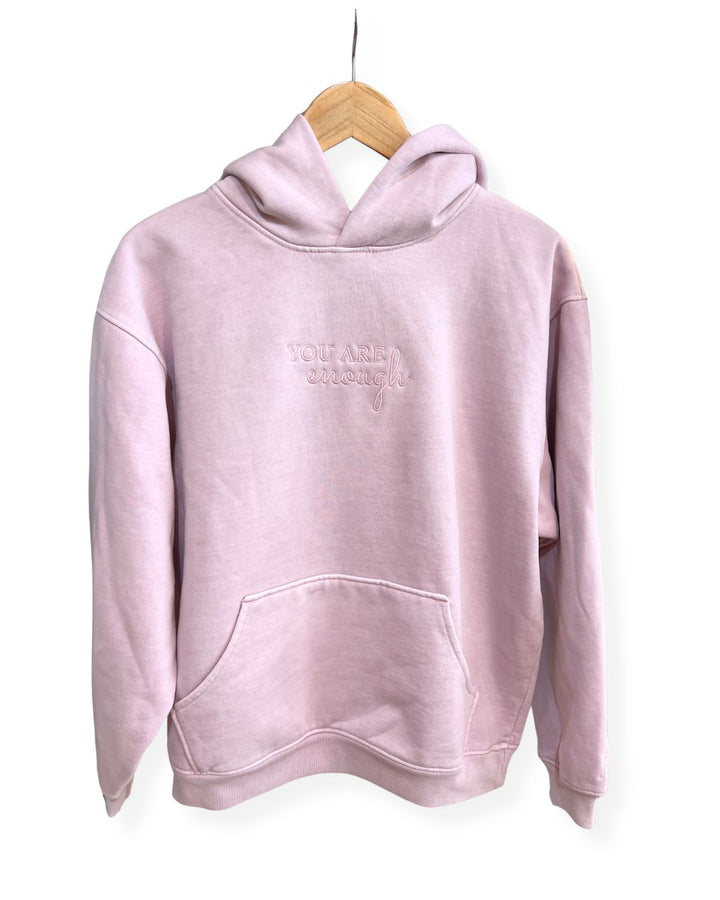 Front view of a light pink, oversized fleece janegee hoodie hanging on a wooden hanger, featuring a kangaroo pocket and "YOU ARE enough" embroidered centered on the chest.