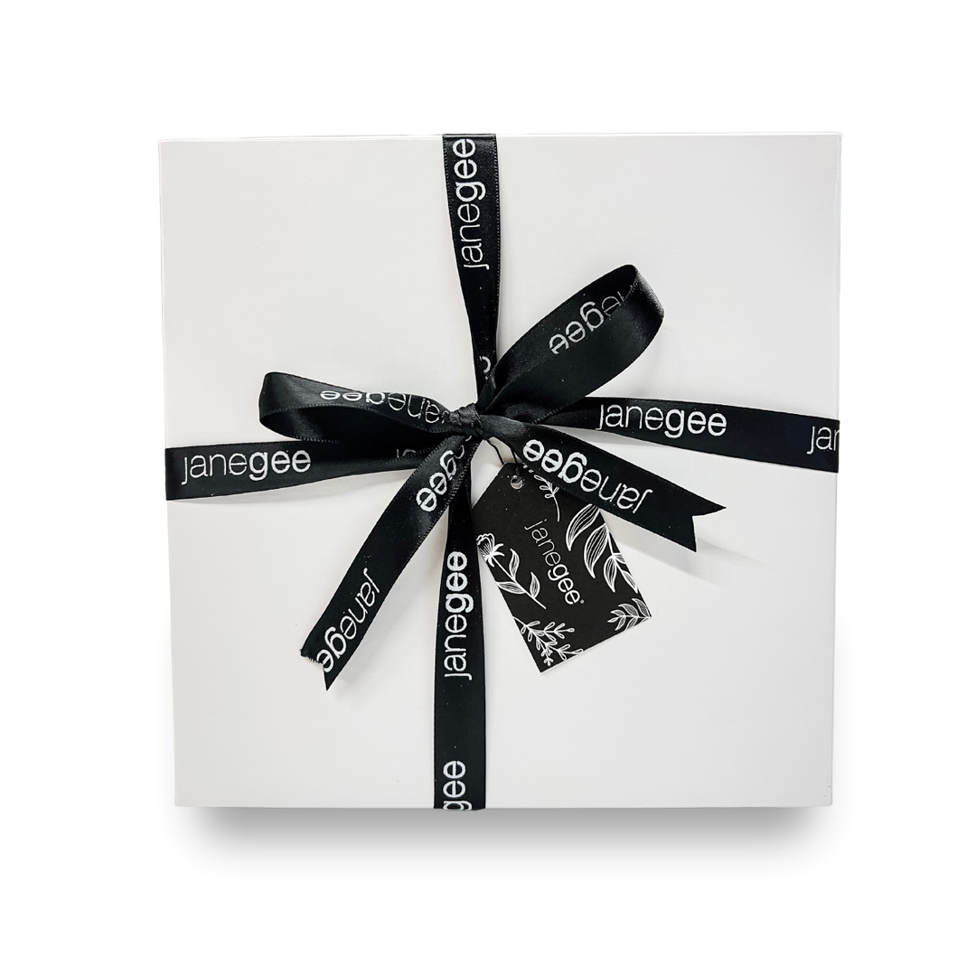 Large white janegee gift box wrapped with black branded ribbon and a botanical gift tag on a soft neutral background.