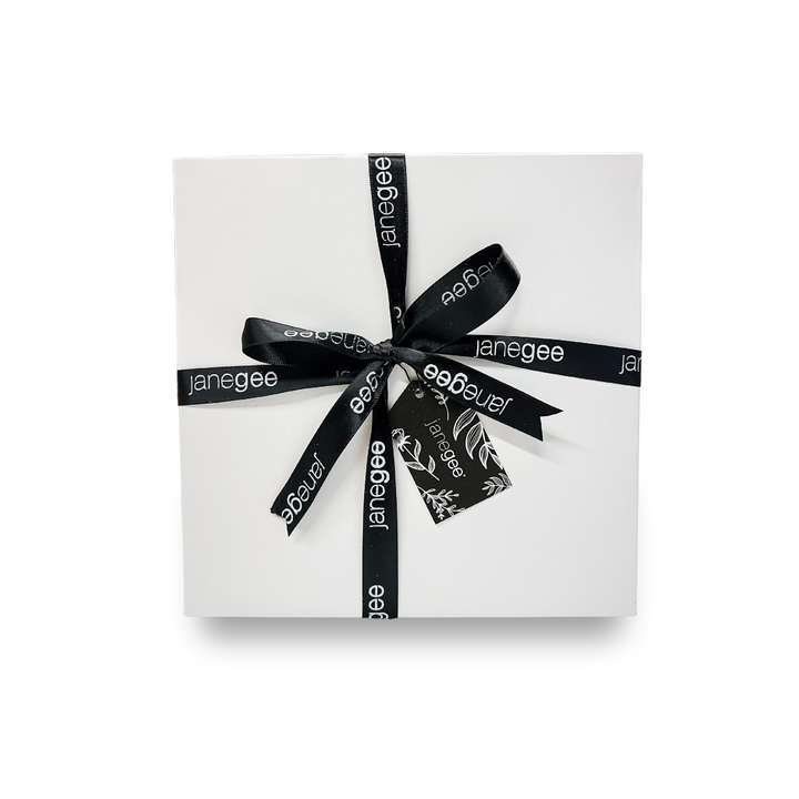 Medium white janegee gift box wrapped with black branded ribbon and a botanical gift tag on a soft neutral background.
