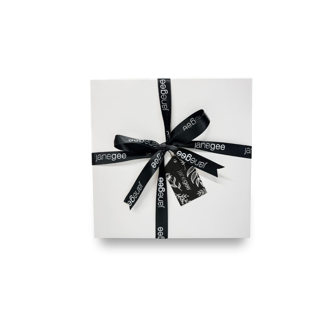 Small white janegee gift box wrapped with black branded ribbon and a botanical gift tag on a soft neutral background.
